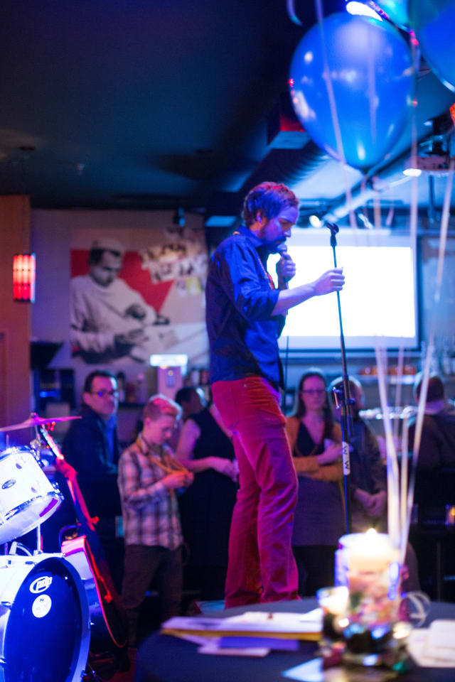 Man speaking into a microphone on stage at Love Bomb 2017 event, audience members in the background, colorful balloons and drum set visible, highlighting community engagement for My Voice Music nonprofit.